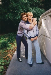 Two men posing outdoors with one man embracing the other from behind wearing patterned shirt and track jacket