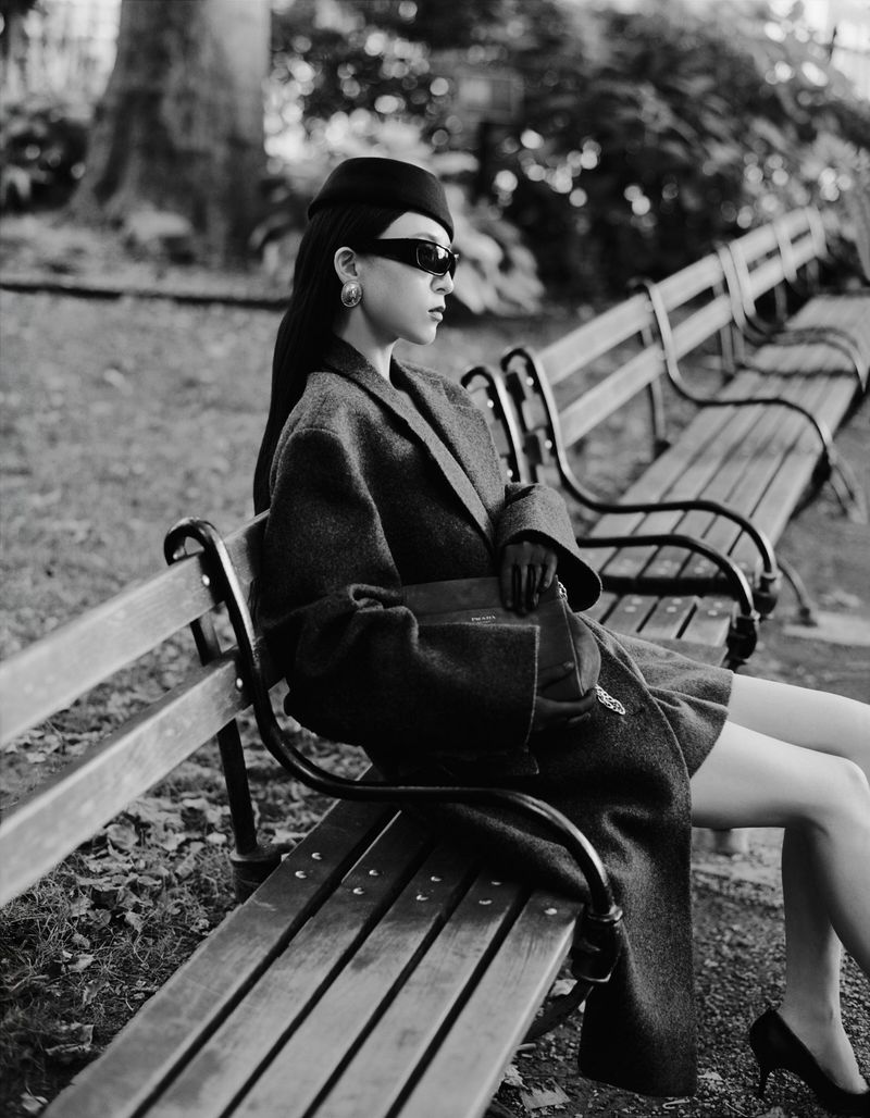 Woman in wool coat and hat sitting on a park bench in an urban environment, styled with sunglasses and black high heels.