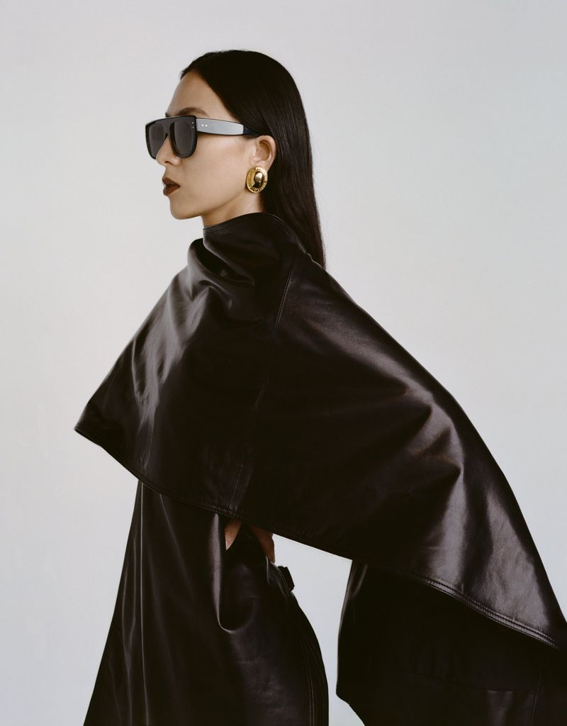 Asian woman in a black leather outfit and sunglasses poses in front of a light gray background in a studio for a fashion editorial.