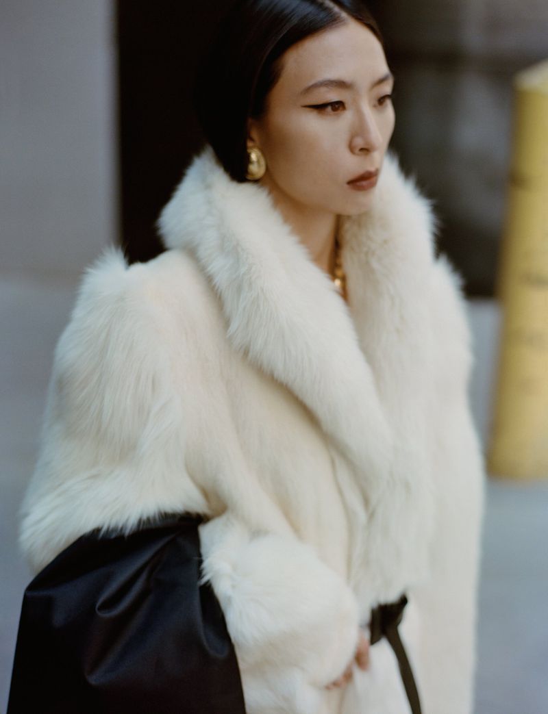 Elegant Asian woman wearing a white fur coat with a black belt in a sophisticated urban environment, showcasing fashion and style.