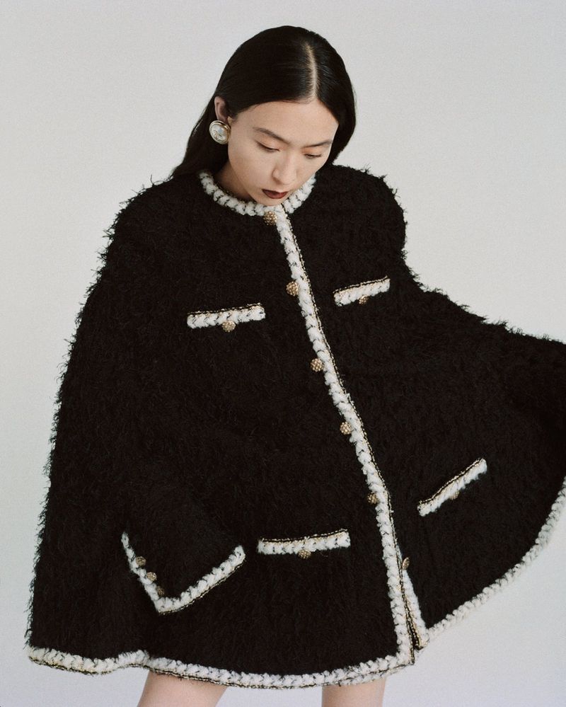 Fashionable Asian woman wearing a chic black tweed jacket with white trim, posing in a brightly lit studio against a white backdrop.