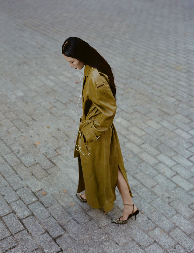 Overhead medium shot of a woman with long dark hair wearing a trench coat and high heels standing on a gray brick road.