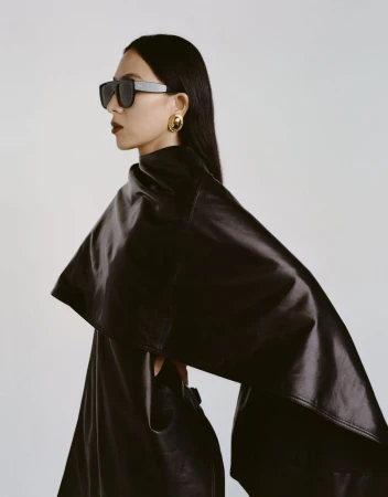 Model in dark sunglasses and black leather cape wearing gold earrings against a light grey studio background.