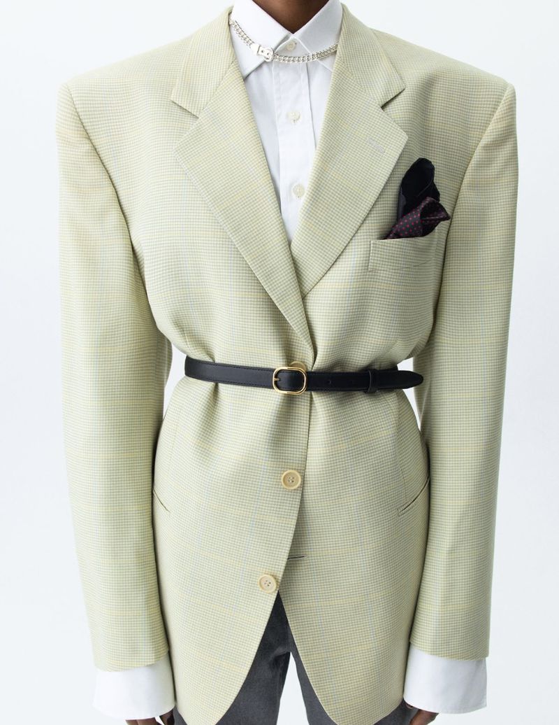 Oversized blazer outfit featuring a houndstooth pattern, cinched with a black belt, accessorized with a silver necklace, styled indoors.
