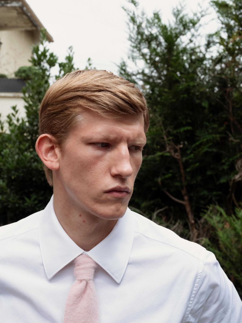 Portrait of a blond adult male in a white collared shirt and pink tie posing outdoors with green foliage in the background.