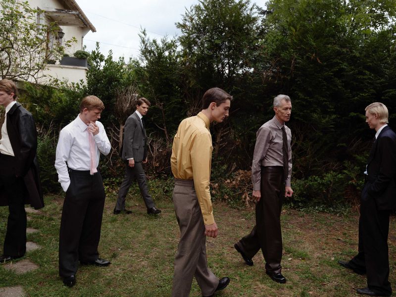 Group of men in suits standing and walking on a lawn outside a building with green trees and bushes in the background.