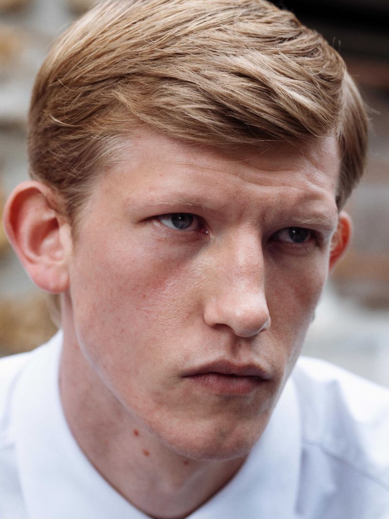 Close-up portrait of a blonde-haired adult male wearing a white collared shirt, captured in natural outdoor lighting with a brick wall backdrop.