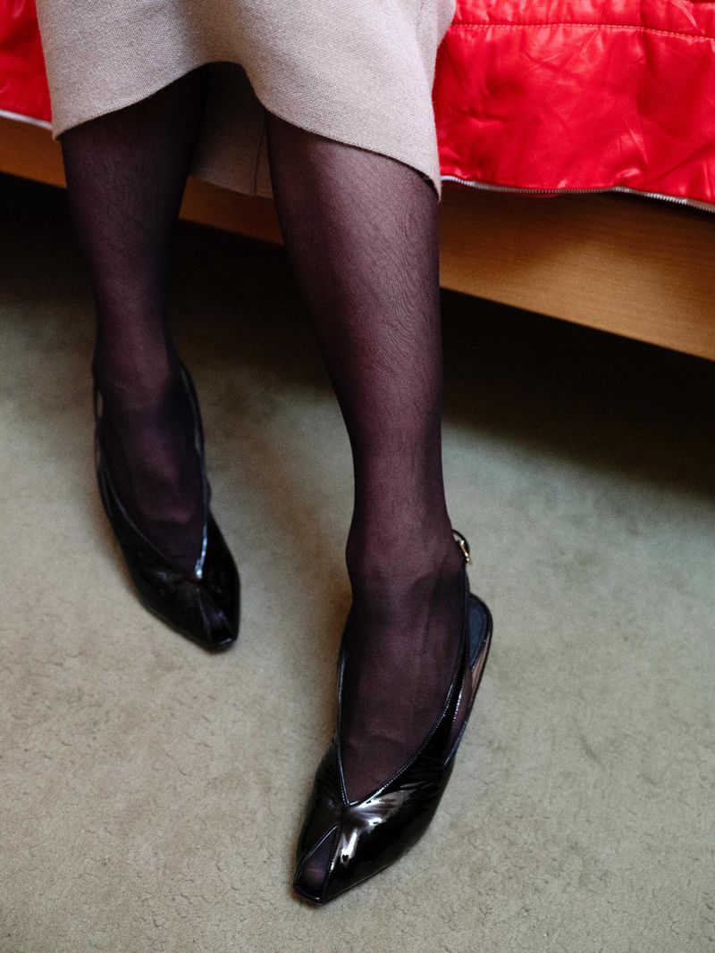 Elegant woman's legs with sheer black stockings and shiny black slingback heels on a neutral carpet, complemented by a tan skirt and red bed.