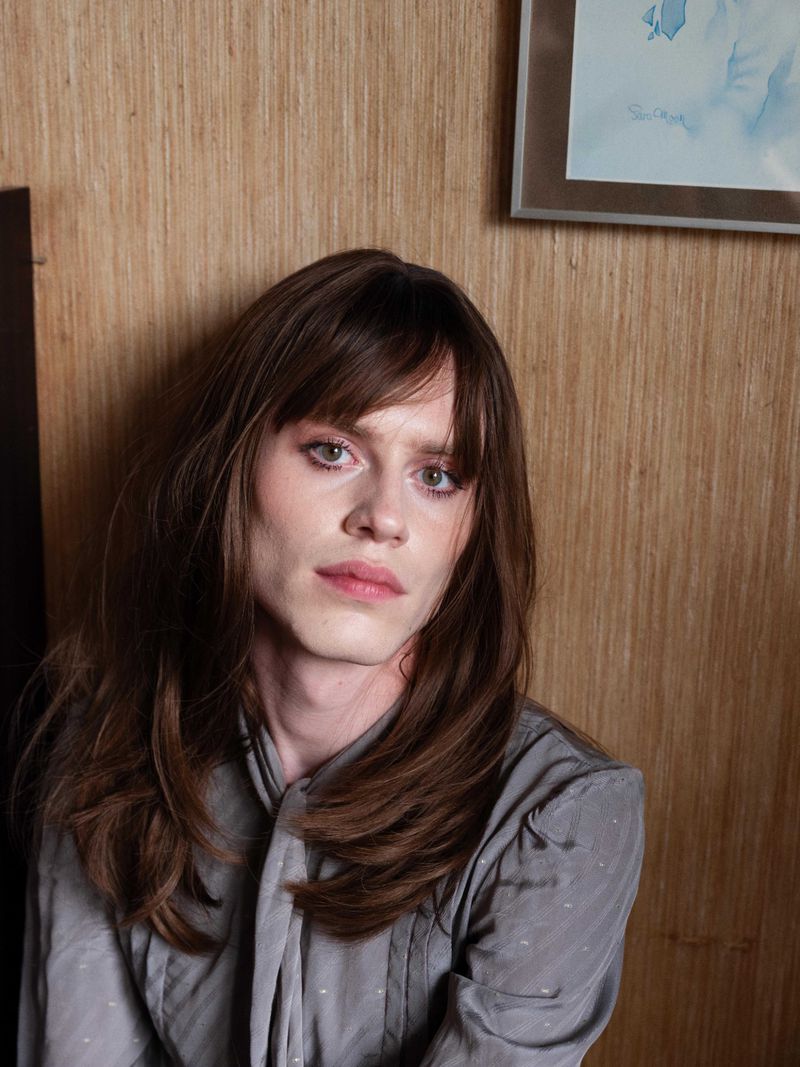 Close-up studio portrait of a young adult with long brown hair and a grey blouse posing with a neutral expression, against a wood-panelled wall.