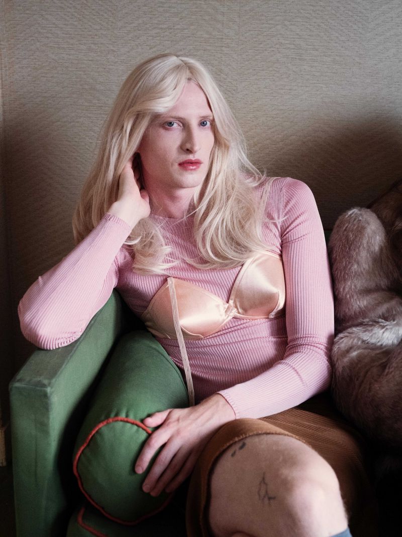 Portrait of a fair-skinned person with long blonde hair in a pink top and bra sitting on a green velvet couch in an indoor setting.