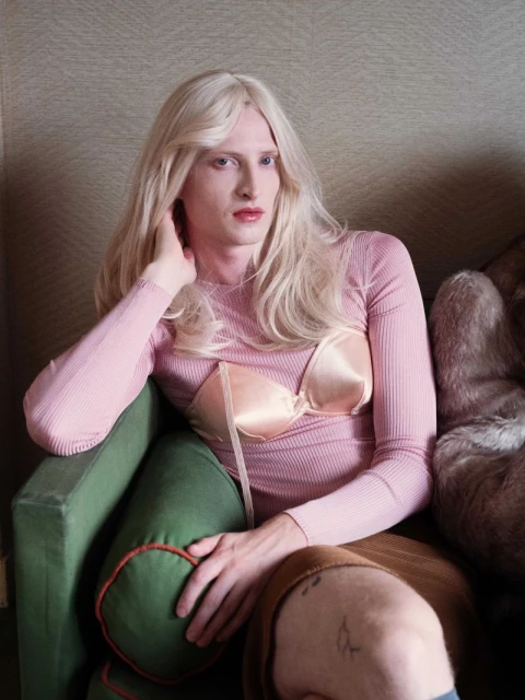 Portrait of a fair-skinned person with long blonde hair in a pink top and bra sitting on a green velvet couch in an indoor setting.