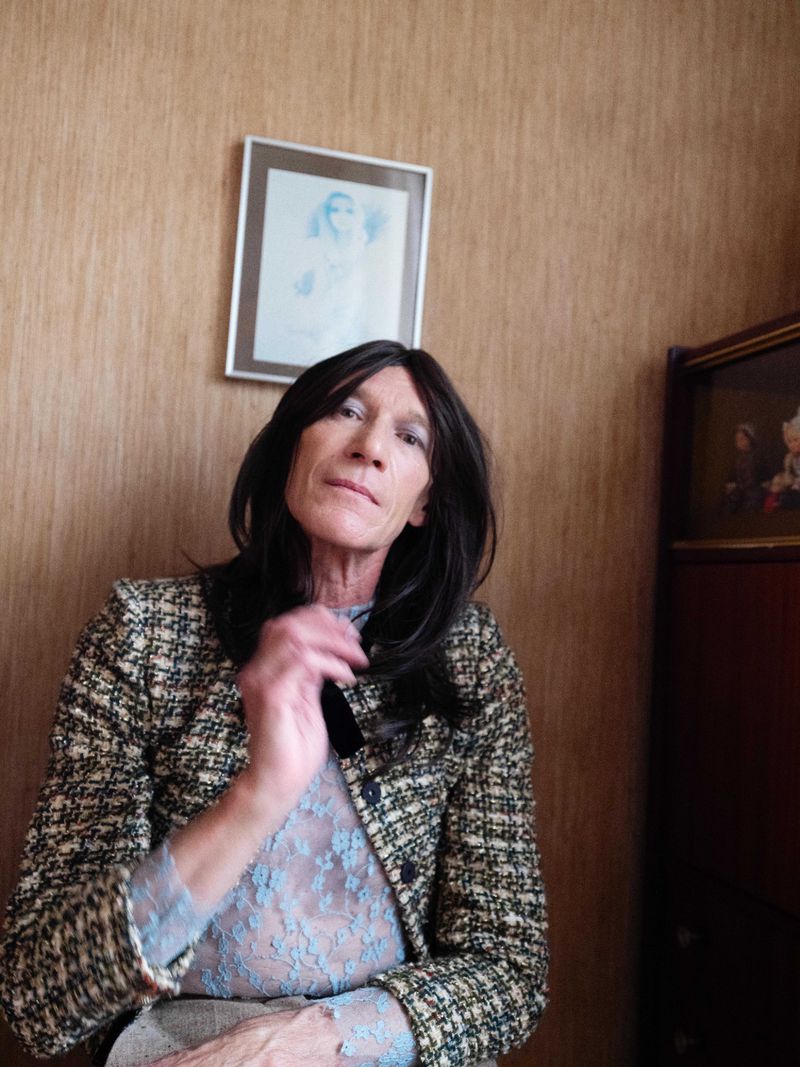 Adult female with dark hair, light complexion, wearing a patterned jacket and a lacy top, sitting indoors in front of a beige wall.