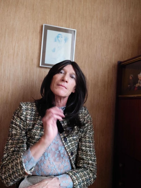 Adult female with dark hair, light complexion, wearing a patterned jacket and a lacy top, sitting indoors in front of a beige wall.