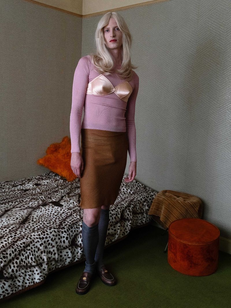 Full-length portrait of a slender adult with blonde hair wearing a pink top, bra, and brown skirt in a bedroom-like setting.
