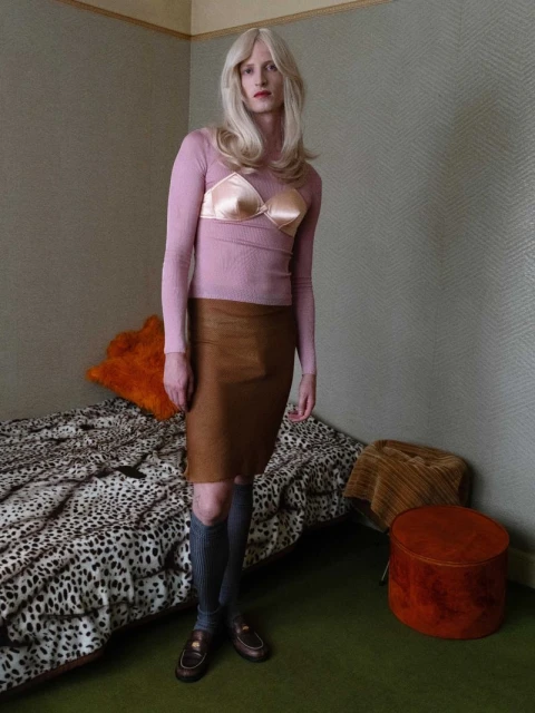 Full-length portrait of a slender adult with blonde hair wearing a pink top, bra, and brown skirt in a bedroom-like setting.