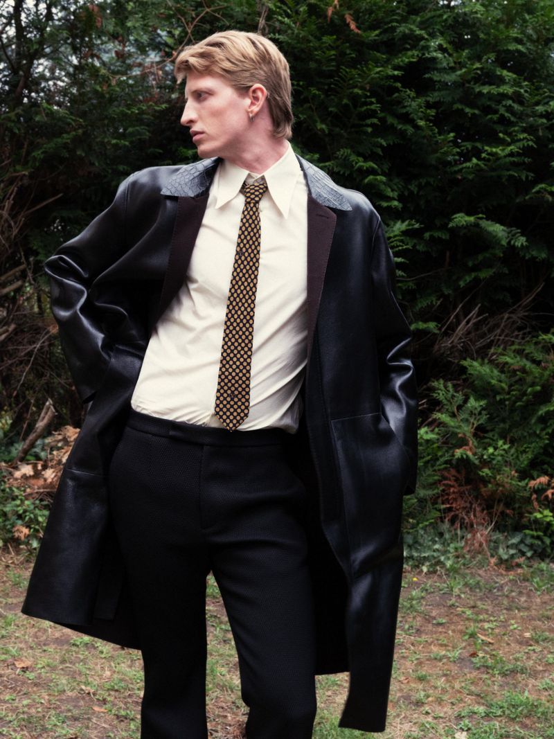 Blonde male model in an outdoor fashion shoot, wearing a black leather trench coat, light collared shirt, and patterned tie.