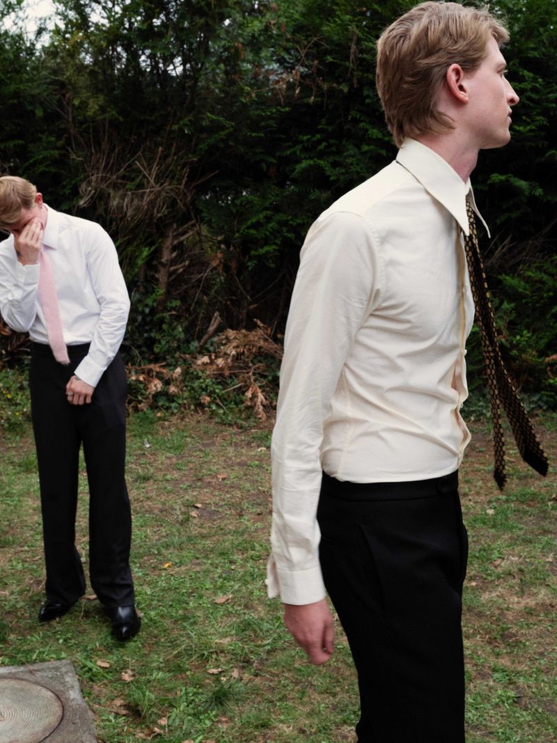 Two adult men in white dress shirts and dark pants stand in a green outdoor environment. One man looks off to the side while the other has head down.