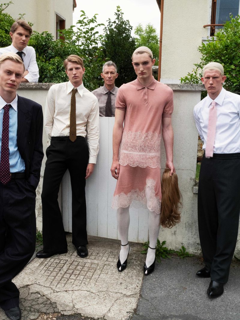 A group of young adult men stand outside in a variety of outfits including suits and a pink dress with lace, holding a wig.