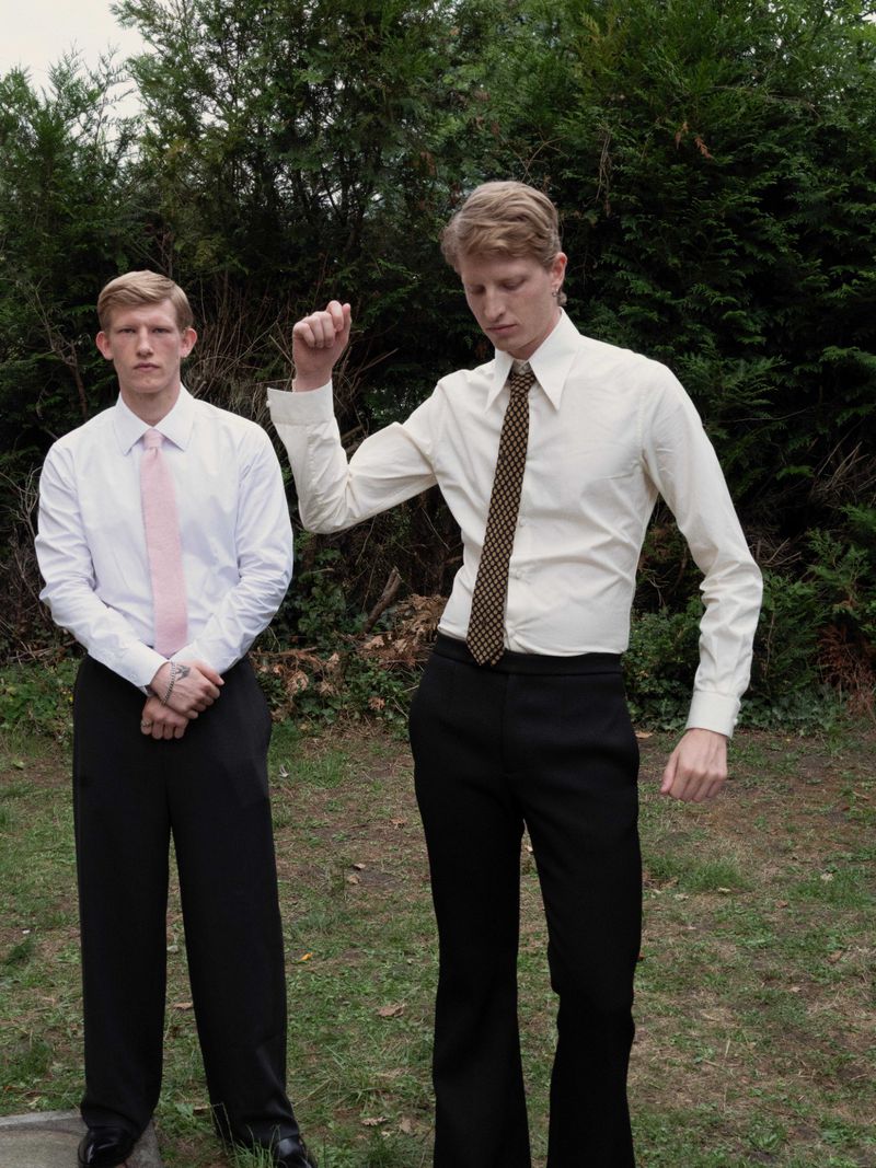 Two fair-skinned adult men in formal attire stand outside; one man raises arm, both wear dress shirts and ties in a natural outdoor setting.