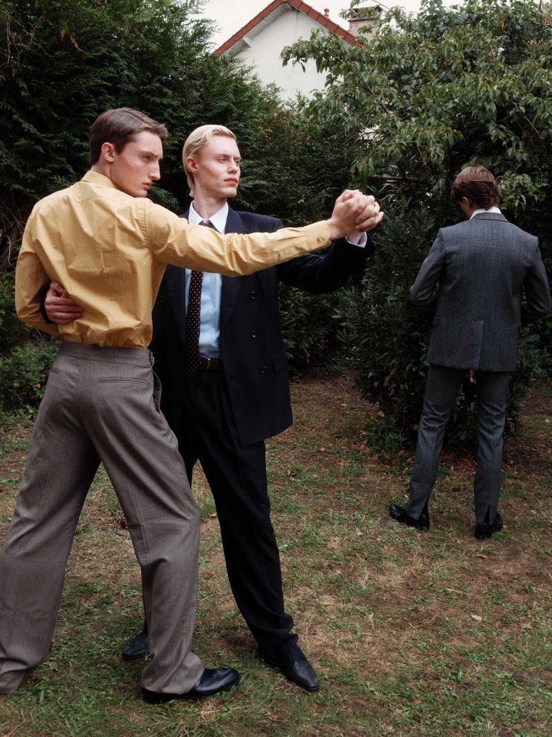 Three men outdoors: two in suits dancing in the foreground, one in a suit out of focus in the background next to a hedge.