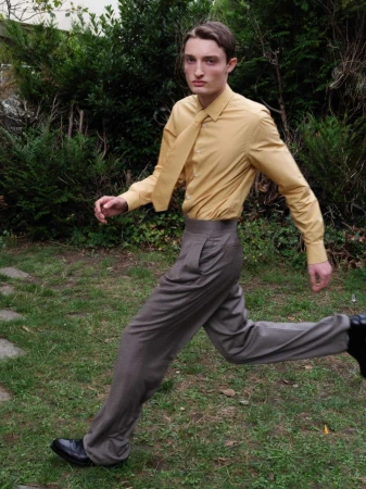 An adult male strides across a grass lawn, dressed in a gold shirt and grey pants, with greenery providing a natural backdrop.