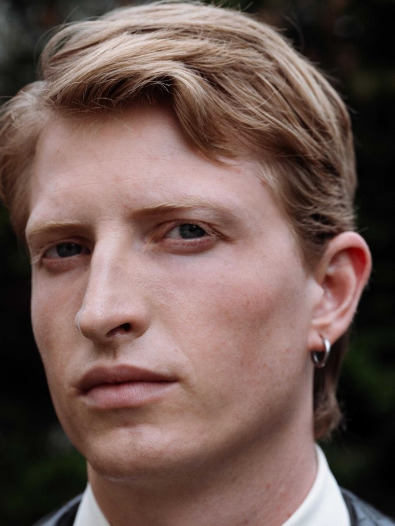 Close-up portrait of a blond man with a serious expression, wearing a collared shirt and dark vest, with a blurred nature background.