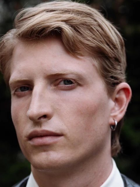 Close-up portrait of a blond man with a serious expression, wearing a collared shirt and dark vest, with a blurred nature background.