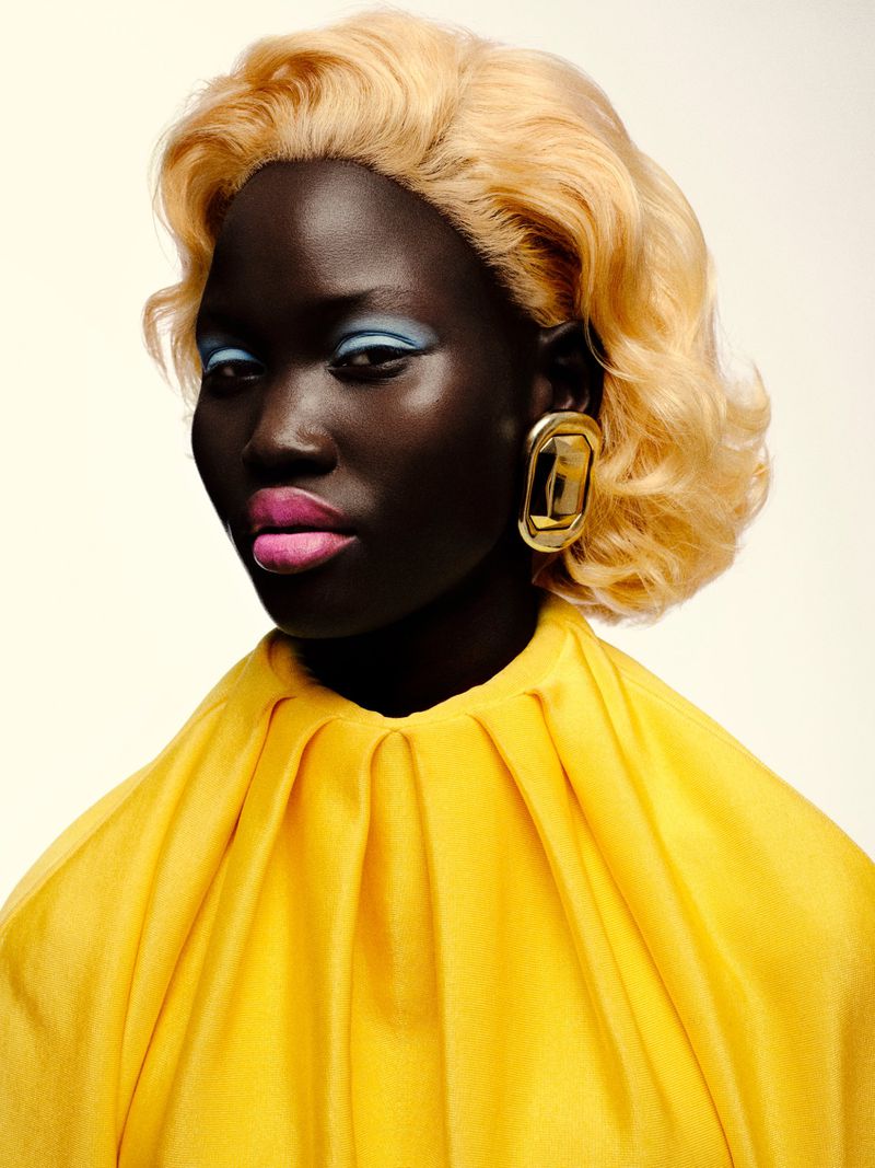 Striking studio portrait of a dark-skinned model with blonde wavy hair, vibrant blue eyeshadow, pink lipstick, and a draped yellow top.