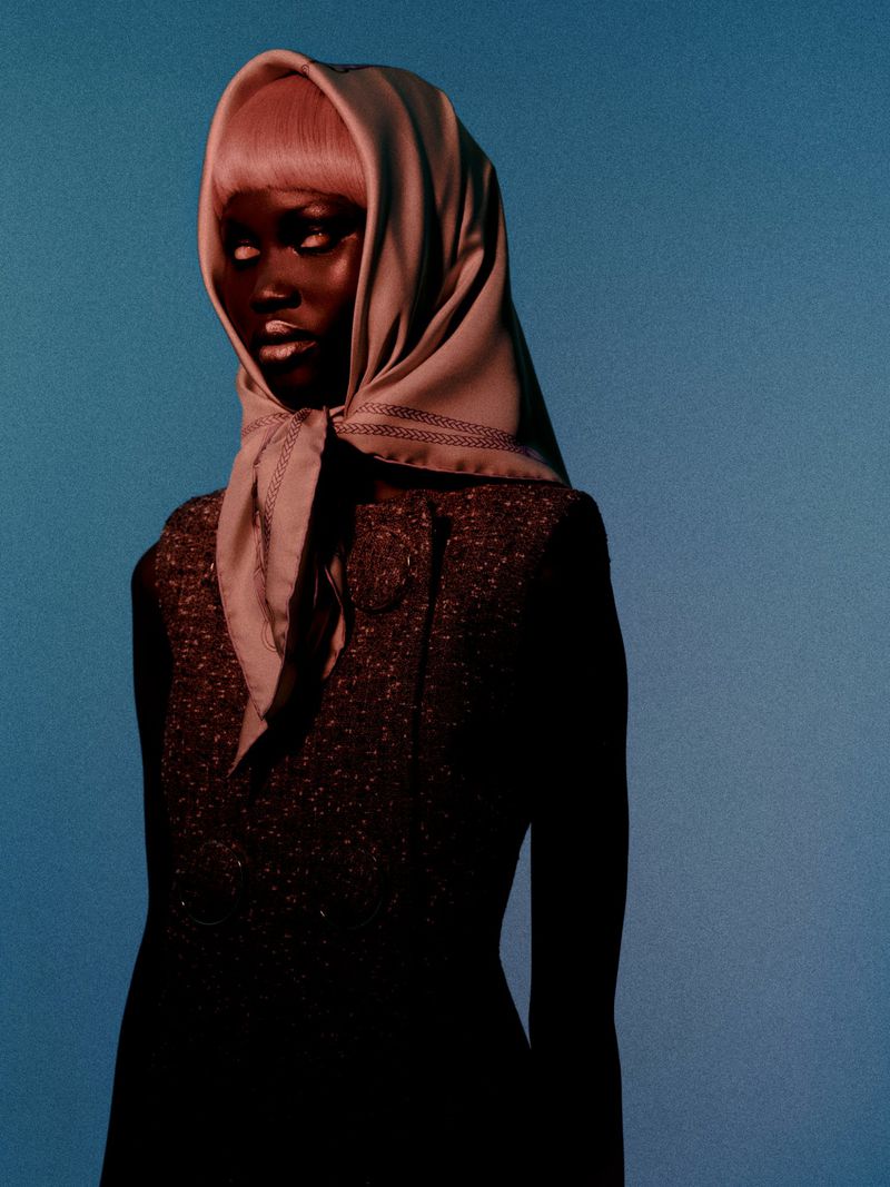 Fashionable woman wearing a headscarf and dark dress posing against a blue backdrop in a studio setting, captured in a medium shot.