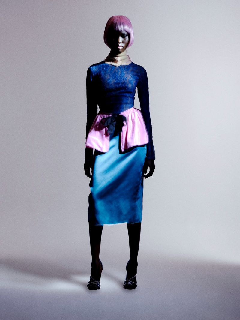 Full-length studio shot of a woman wearing a blue top, pink and blue skirt, and heels against a grey background.
