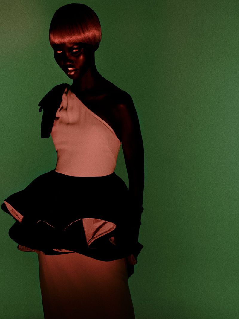 Woman in a one-shoulder dress with a dramatic black ruffle, posing in a studio with a solid green backdrop, fashion portrait.