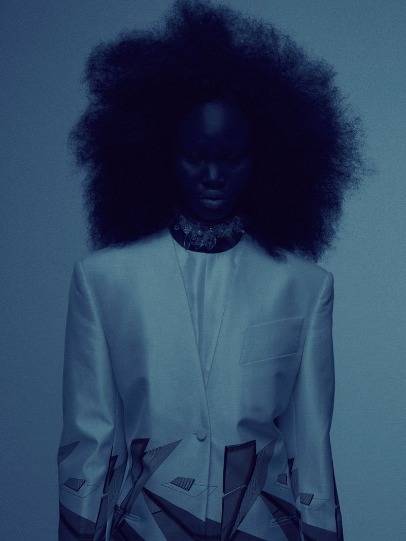 A stylish black model in an off-white blazer with a geometric pattern poses in a studio with a gradient blue background.