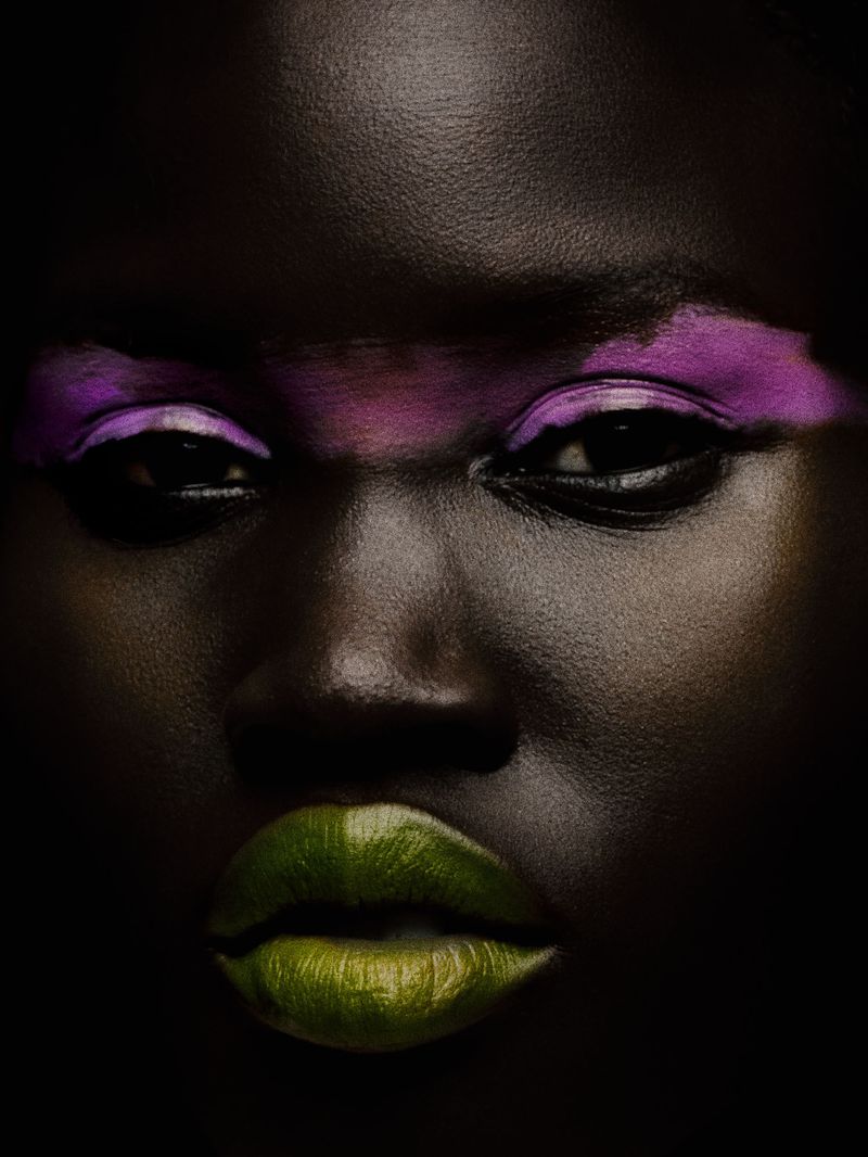Close-up portrait of an adult woman with dark skin, vivid pink eyeshadow, and green lipstick against a dark backdrop.