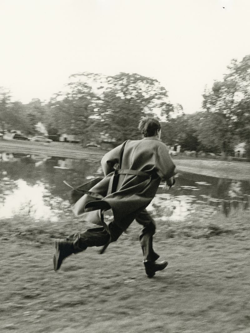 A man in a long belted coat running outdoors near a body of water in a black and white editorial photograph.