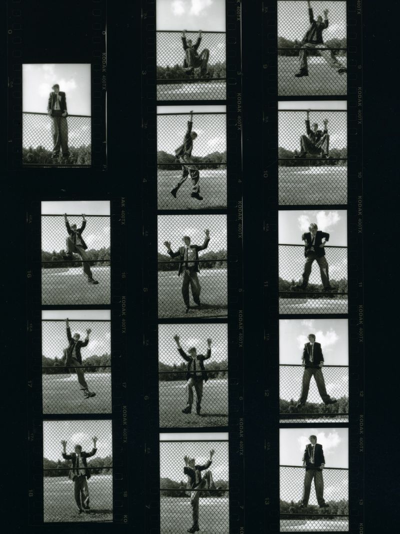 Black and white film strip of a man in a suit, interacting with a chain link fence in various poses against a grassy field and sky.