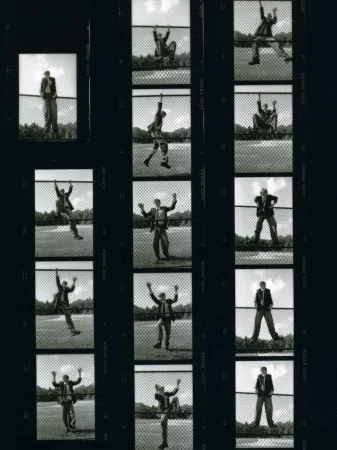 Black and white film strip of a man in a suit, interacting with a chain link fence in various poses against a grassy field and sky.