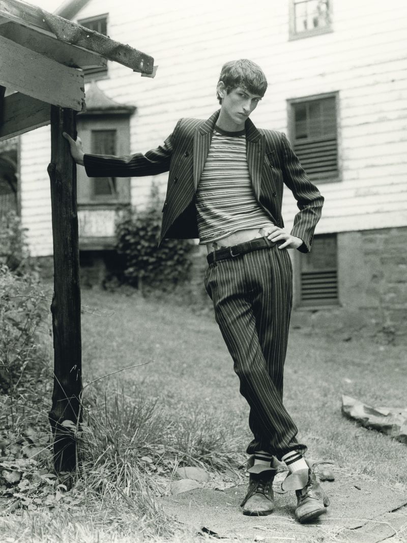 Man poses in pinstripe suit with striped shirt, outside by a weathered building. Black-and-white photography in natural setting with grass.