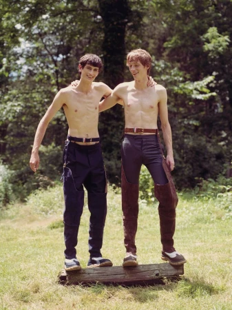 Two shirtless young men standing on a log in a wooded area, wearing pants and shoes, hugging with a grass and tree background.