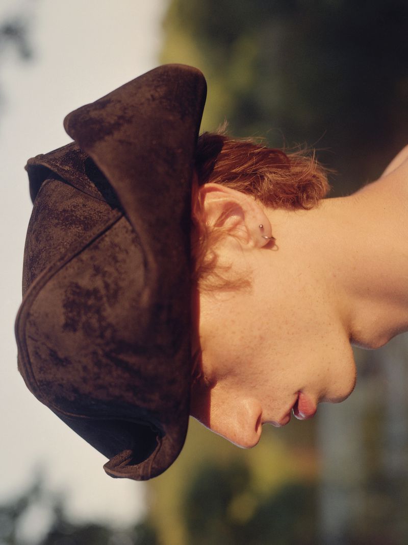 Close-up of a person wearing a historical-style dark brown hat, their face partly shadowed against a blurred green and light-colored background.