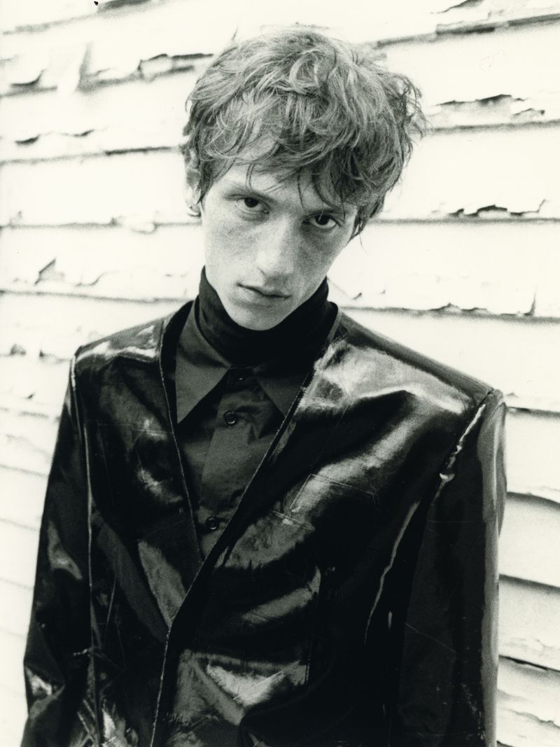 Young man wearing a dark glossy jacket against a weathered white wooden wall in a black and white fashion portrait.