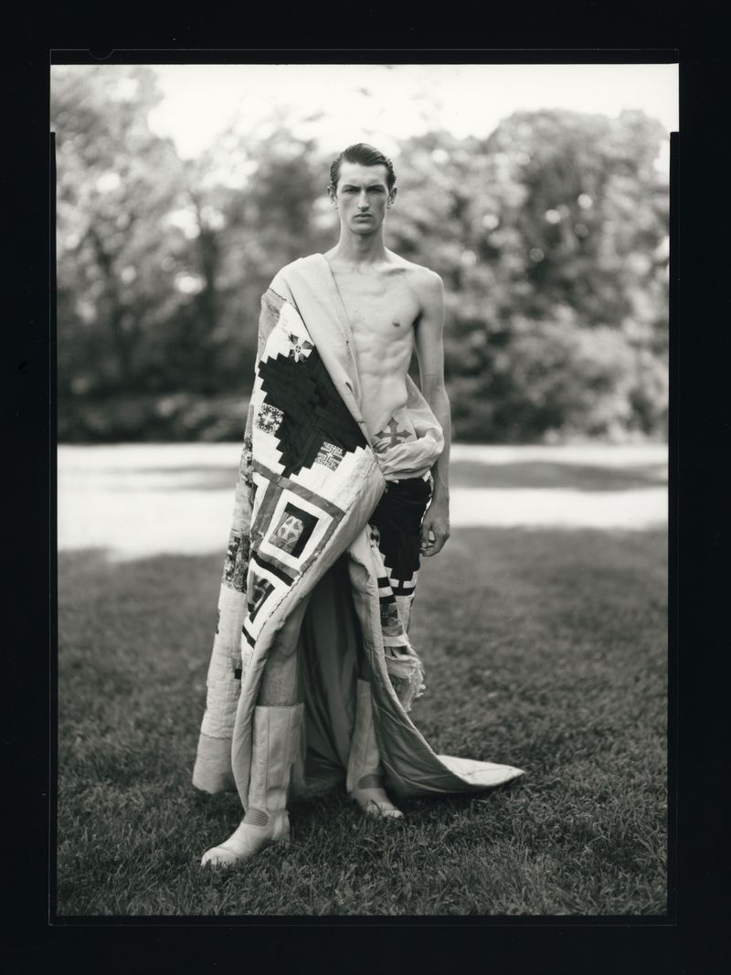 Monochrome portrait of a slender man draped in a patchwork quilt, standing barefoot on grass in a sunlit outdoor setting.