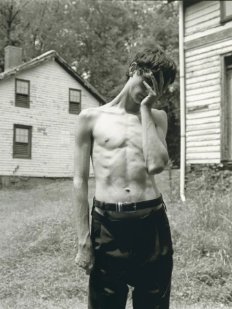 Shirtless young man wearing dark trousers standing outdoors in front of a white clapboard house.