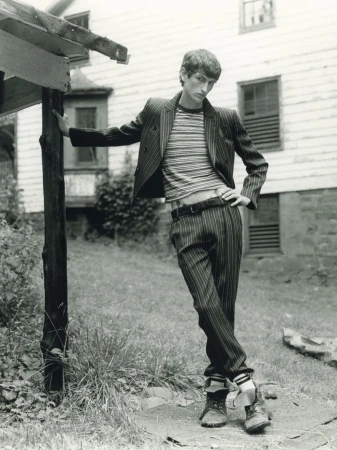 Monochrome portrait of a male model leaning against a wooden post outside a light-colored house in a striped suit.