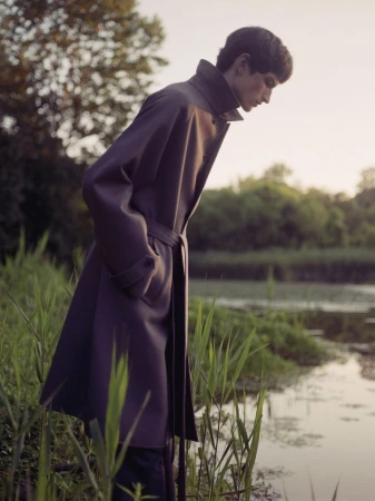 Model wearing a dark belted coat standing by a calm body of water with tall green reeds in the foreground.