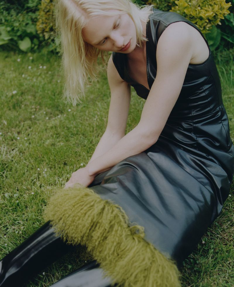 Blonde woman in black dress sits on green grass with fur trim on black boots in a sunny outdoor setting.