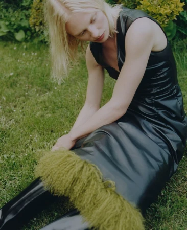 Blonde model wearing a black faux leather dress seated on a lush green lawn for Puss Puss Magazine editorial photography.