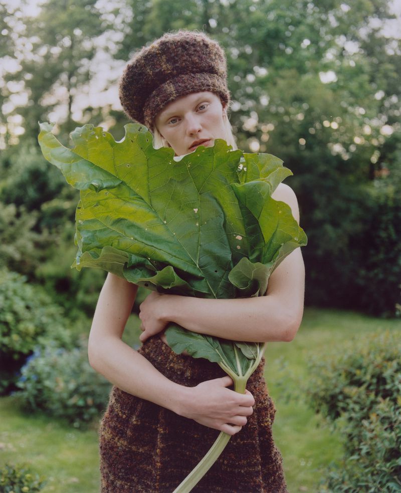 Blonde woman in brown hat and woven dress holds large green leaves in an outdoor garden setting with green foliage.