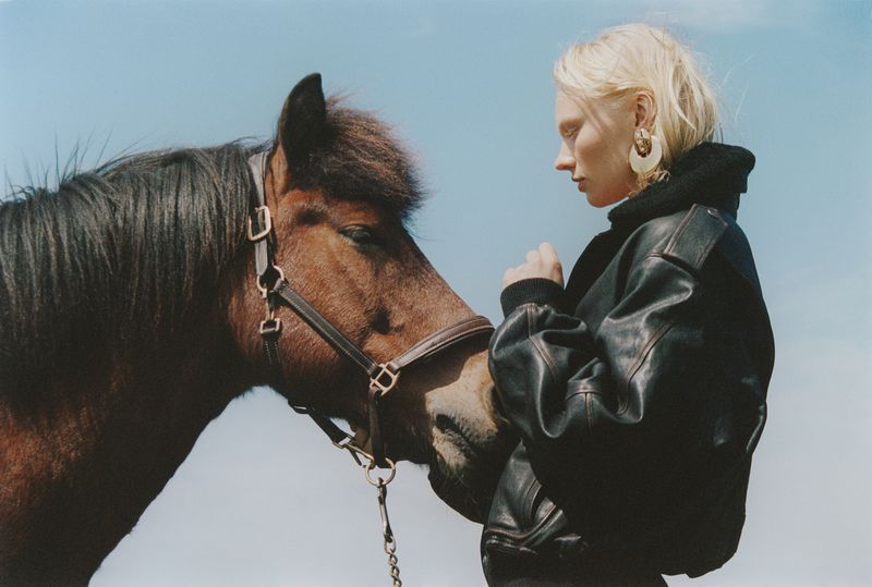 A young blonde woman in a black leather jacket stands outdoors in daylight, nuzzling a brown horse against a pale blue sky backdrop.