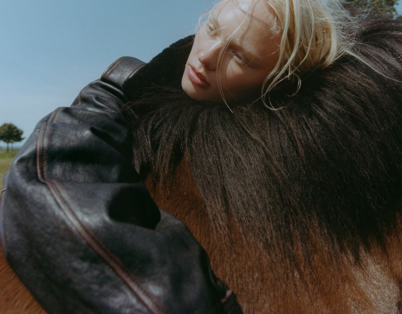 Blonde model wearing a dark leather jacket resting against a horse outdoors under a clear blue sky