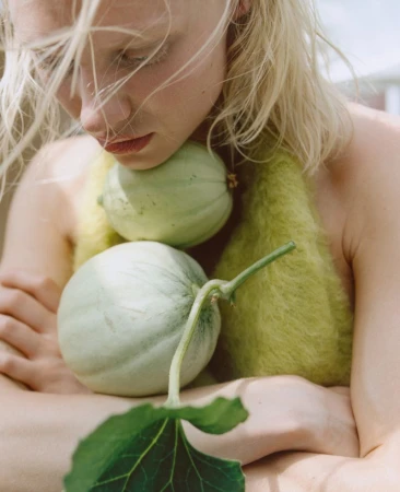 Young woman with blonde hair poses intimately holding two light green melons in her arms, set against an outdoor background.